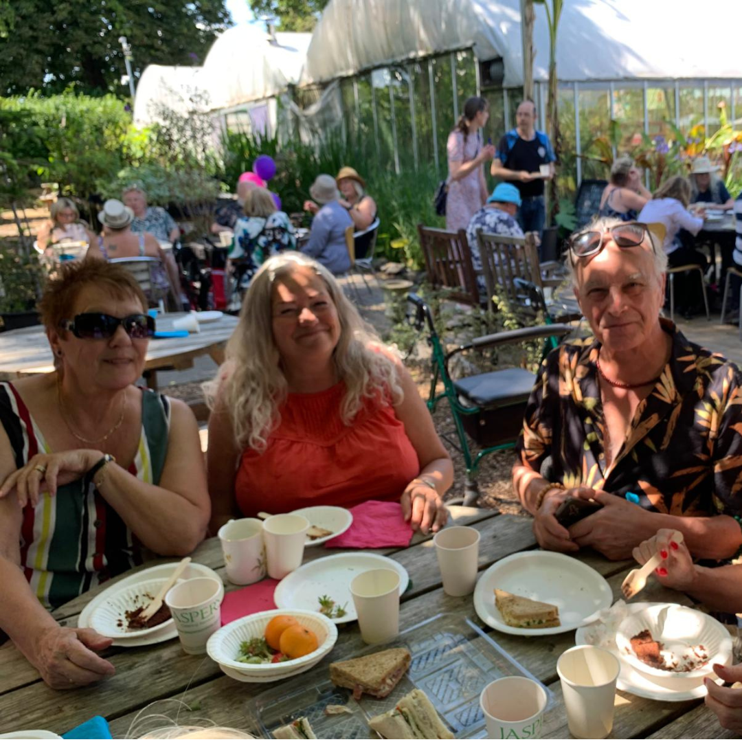 3 guests sat at table smiling, other groups, plants and polytunnels in background