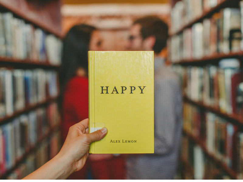 Book entitled 'HAPPY' held up with male and female gazing into each others eyes in a library aisle