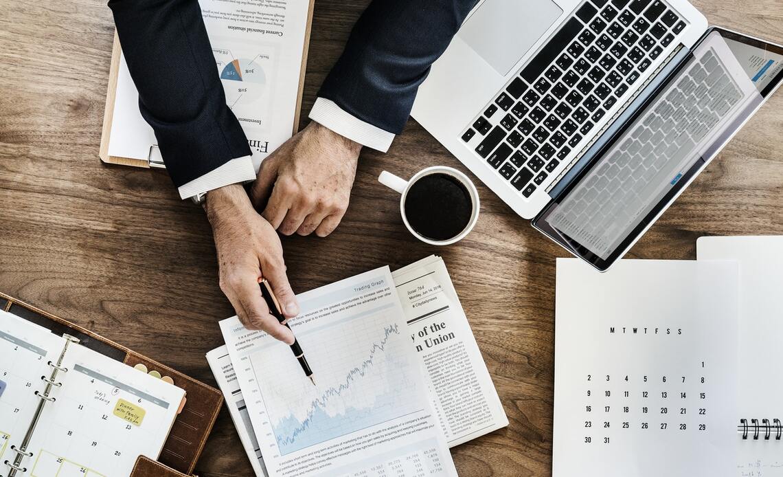 Desk with coffee cup, laptop and man's hands pointing to charts and graphs