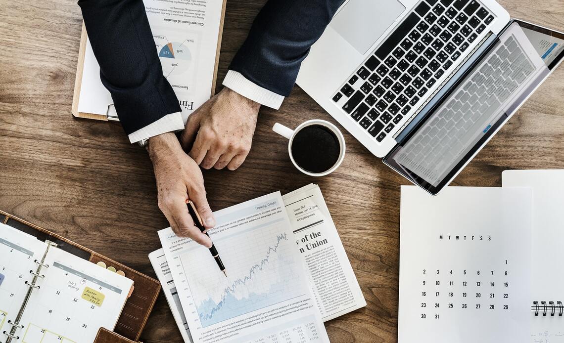 Desk with coffee cup, laptop and man's hands pointing to charts and graphs